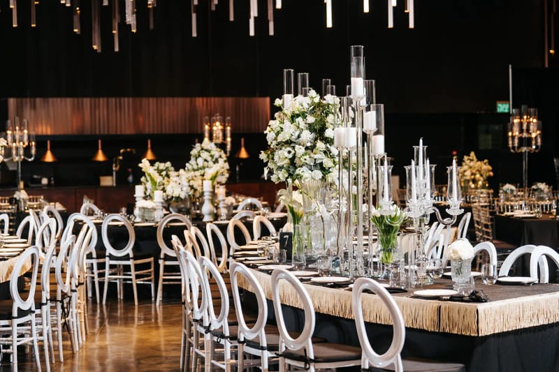 Long fringed-edge table with white chairs, tall glass candle holders and white flowers on a wooden floor