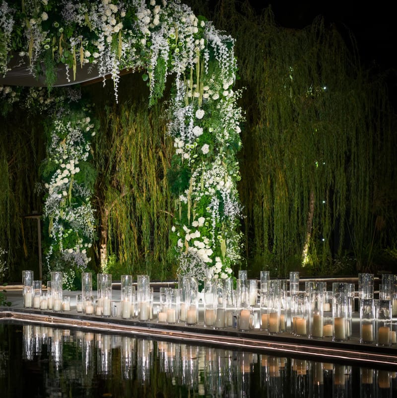 Arch of white flowers and trailing greenery before a wall of willows, with a row of candles at the water's edge