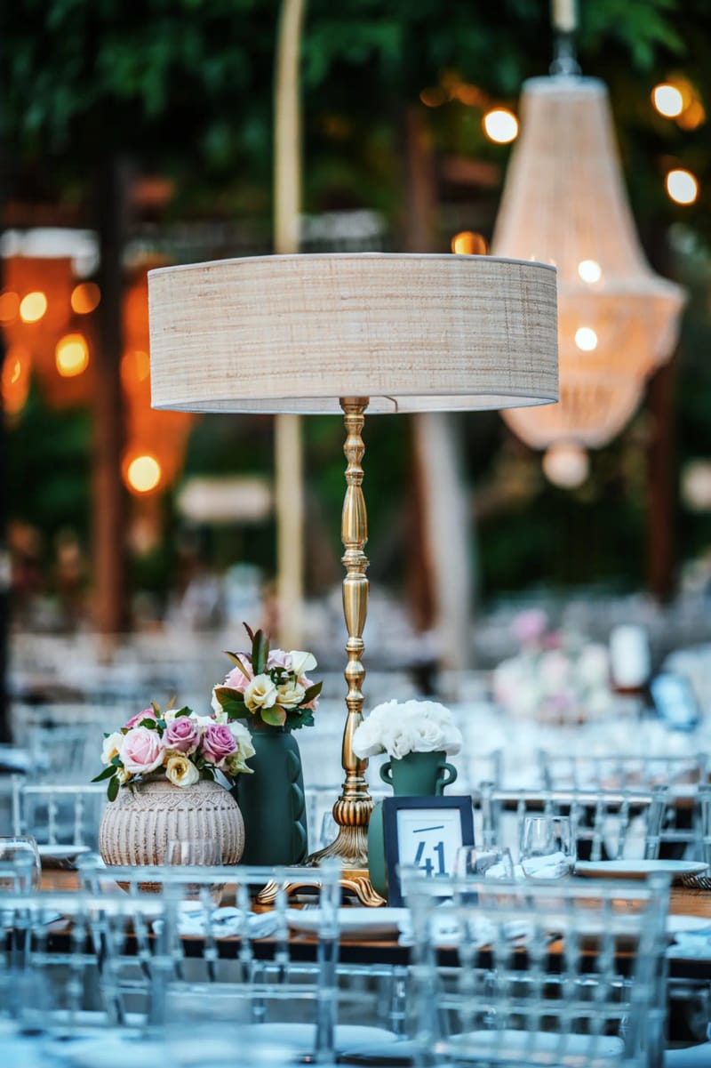 Brass floor lamp with a fabric shade beside a floral arrangement and table number, with a crystal chandelier blurred behind
