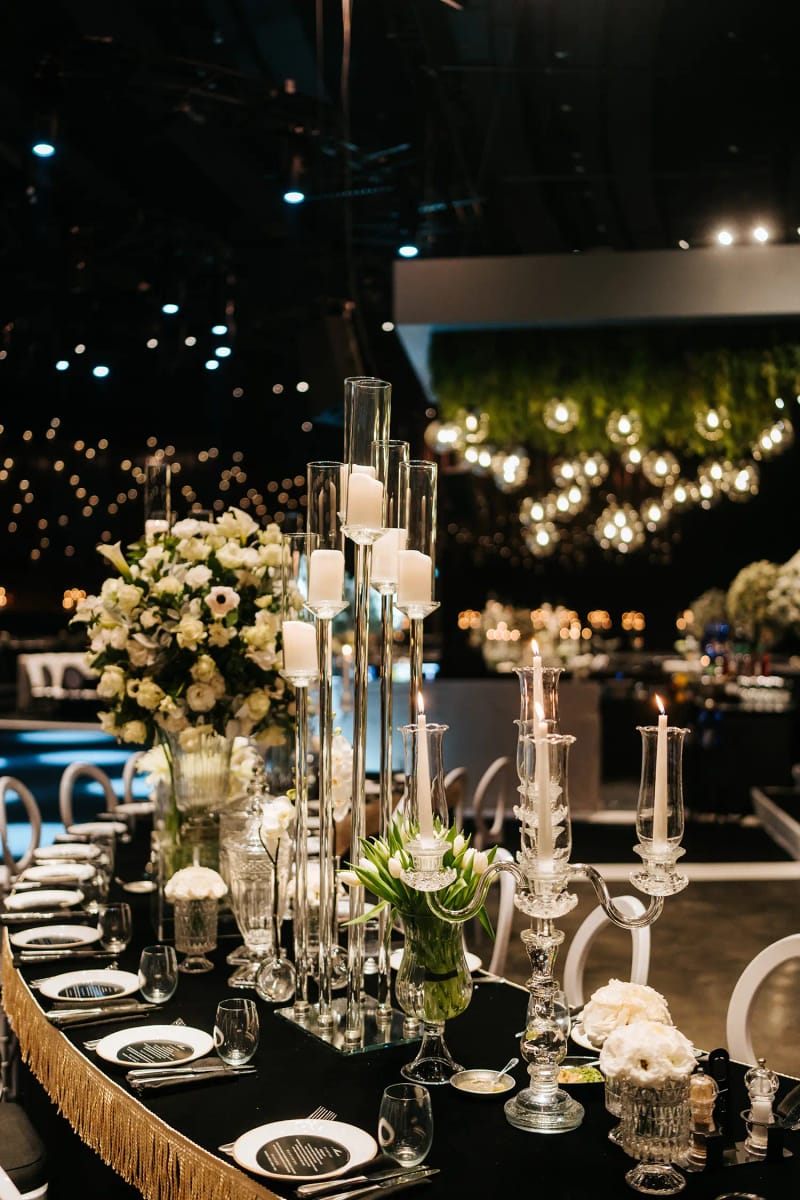 Hanging greenery planter with globe bulbs above a black fringed table with white flowers and glass candle holders