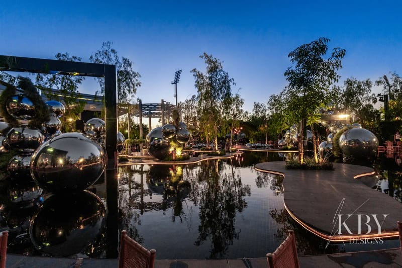 Dark reflective pool with mirror spheres, a mirrored frame and trees at twilight, beside a lit curving walkway