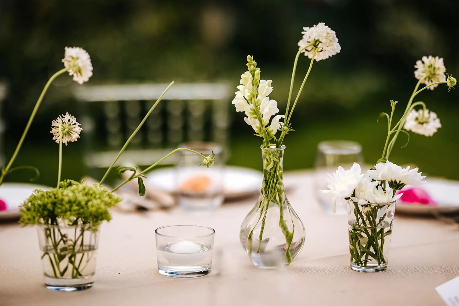 Petits vases avec fleurs blanches et tiges vertes et une bougie dans un verre sur une table dressée