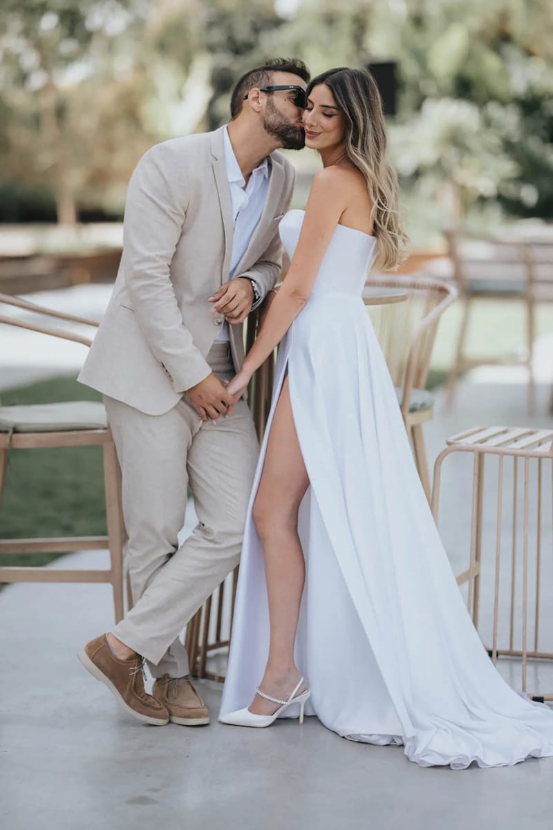A couple embracing as the groom kisses the bride's cheek, beside bar stools on the lawn