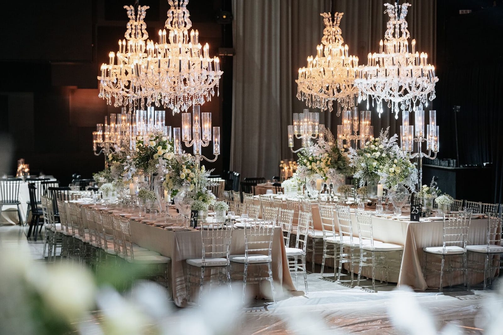 Longue table de banquet aux centres de table floraux, lustres en cristal et chaises transparentes dans une salle aux rideaux sombres