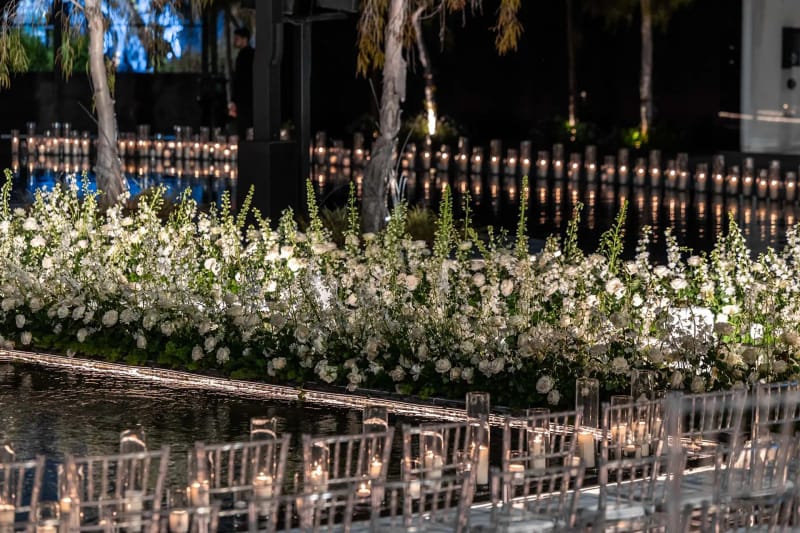 Massif bas de fleurs blanches près d'un bassin avec des bougies au bord de l'eau et des chaises transparentes devant