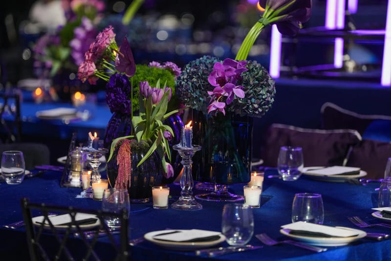 Close-up of a round table with a royal blue cloth, purple flowers and hydrangeas, candles and neon tubes behind
