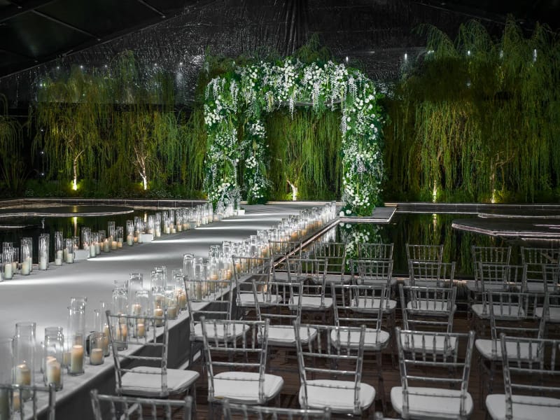 White aisle lined with candles beside dark reflective water at night, with chairs and a floral arch before a wall of willows
