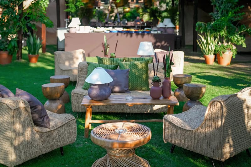 Woven rattan armchairs and a wooden table with lamps and clay vessels on the lawn, among potted plants