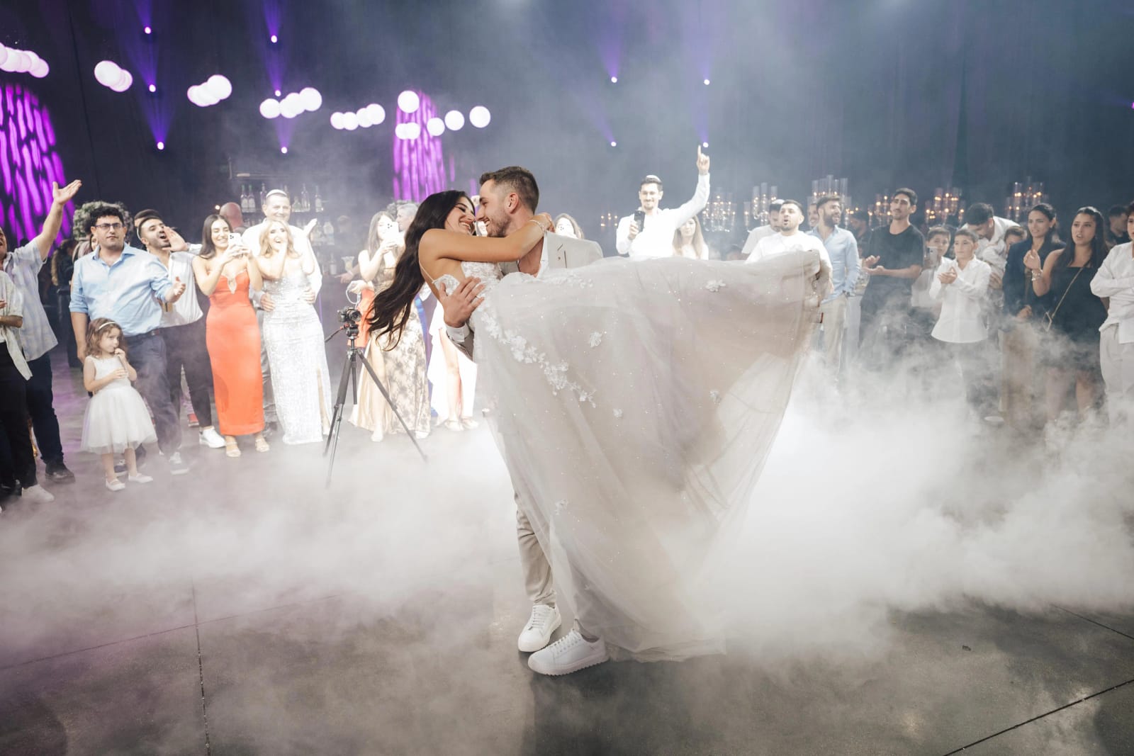 A couple sharing a dipped kiss on a smoke-filled dance floor, surrounded by guests