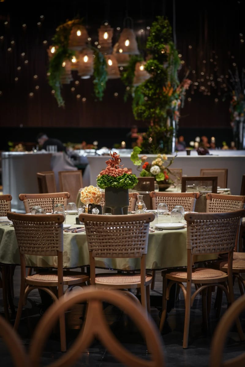 Round table with cane-back chairs and hanging rattan lanterns with greenery above, against a dark wall