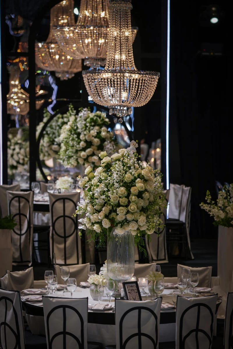 Lustres en cristal au-dessus de tables rondes à hautes compositions blanches et chaises noires, dans une salle sombre