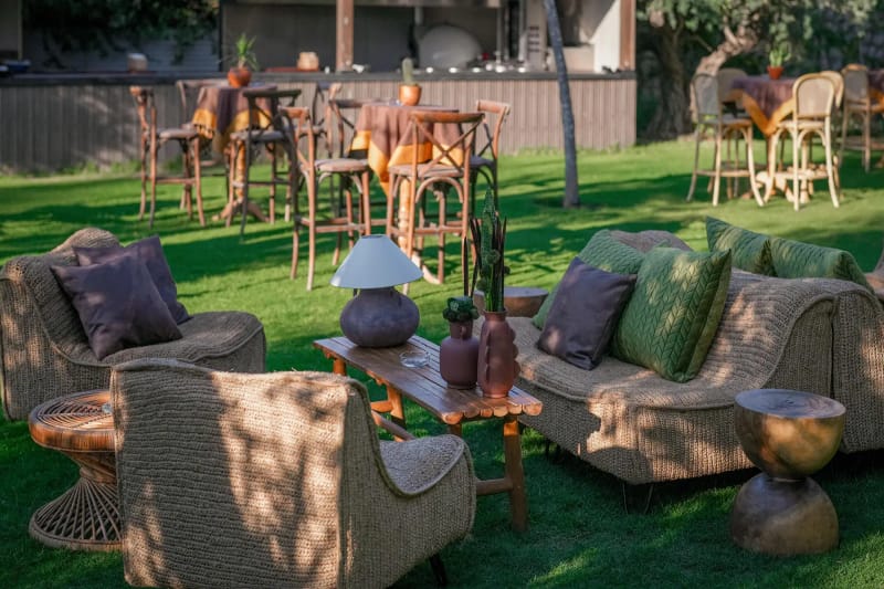 Woven sofa and armchairs with green and plum cushions beside high tables with wooden stools on the lawn