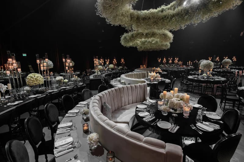 Dark hall with a curved cream banquette, round tables with black chairs and a hanging ring of white flowers above