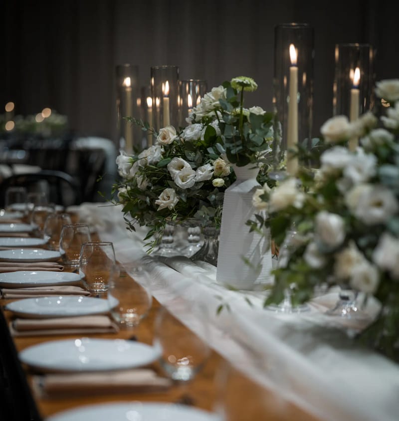 Long wooden table with white plates, a sheer runner, white flower bouquets and candles in glass cylinders