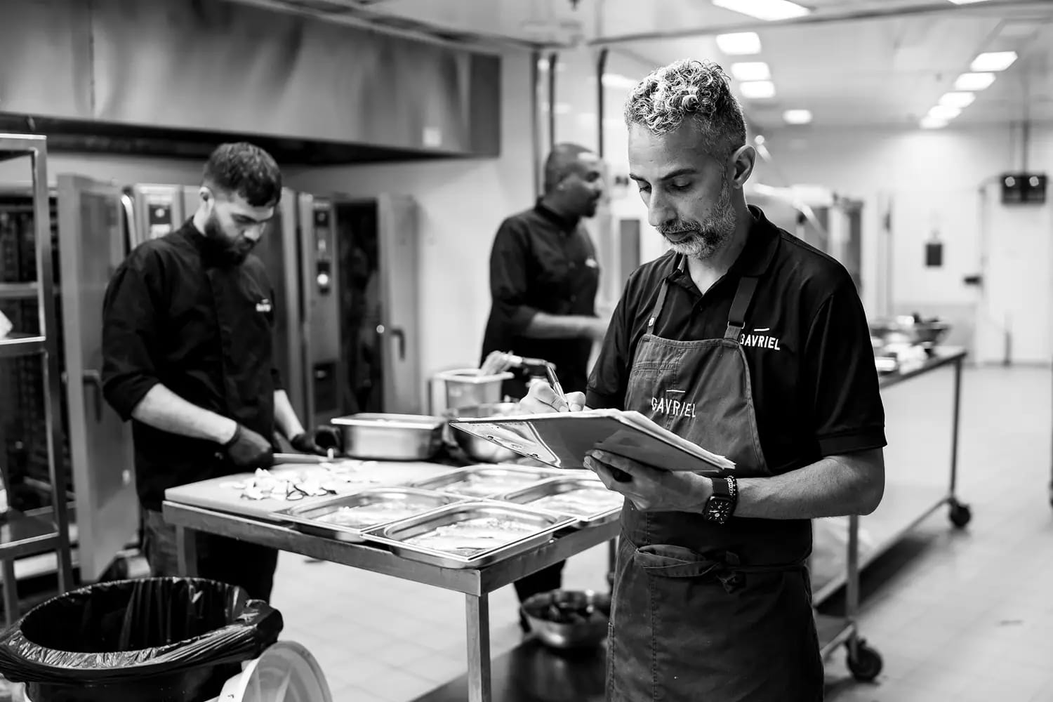 Black and white shot of a chef in an apron reading from a book at a prep table, cooks working in a commercial kitchen behind