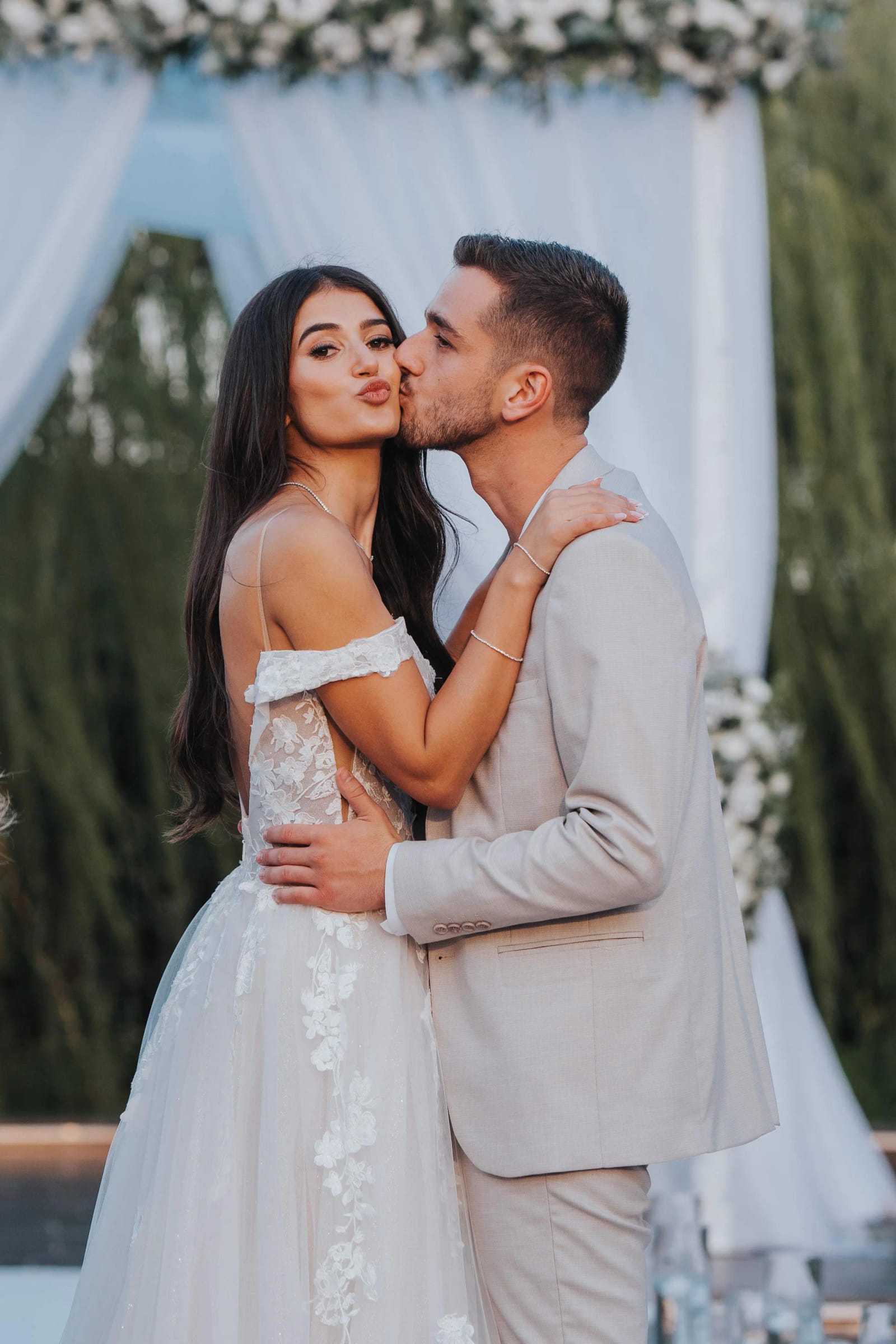A couple embracing, the groom kissing the bride's cheek, in front of white canopy drapes and willow trees
