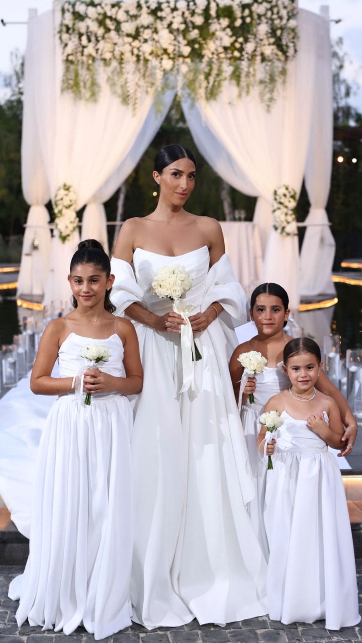 Une mariée en robe blanche entourée de petites filles en robe blanche tenant de petits bouquets sous un dais drapé et fleuri