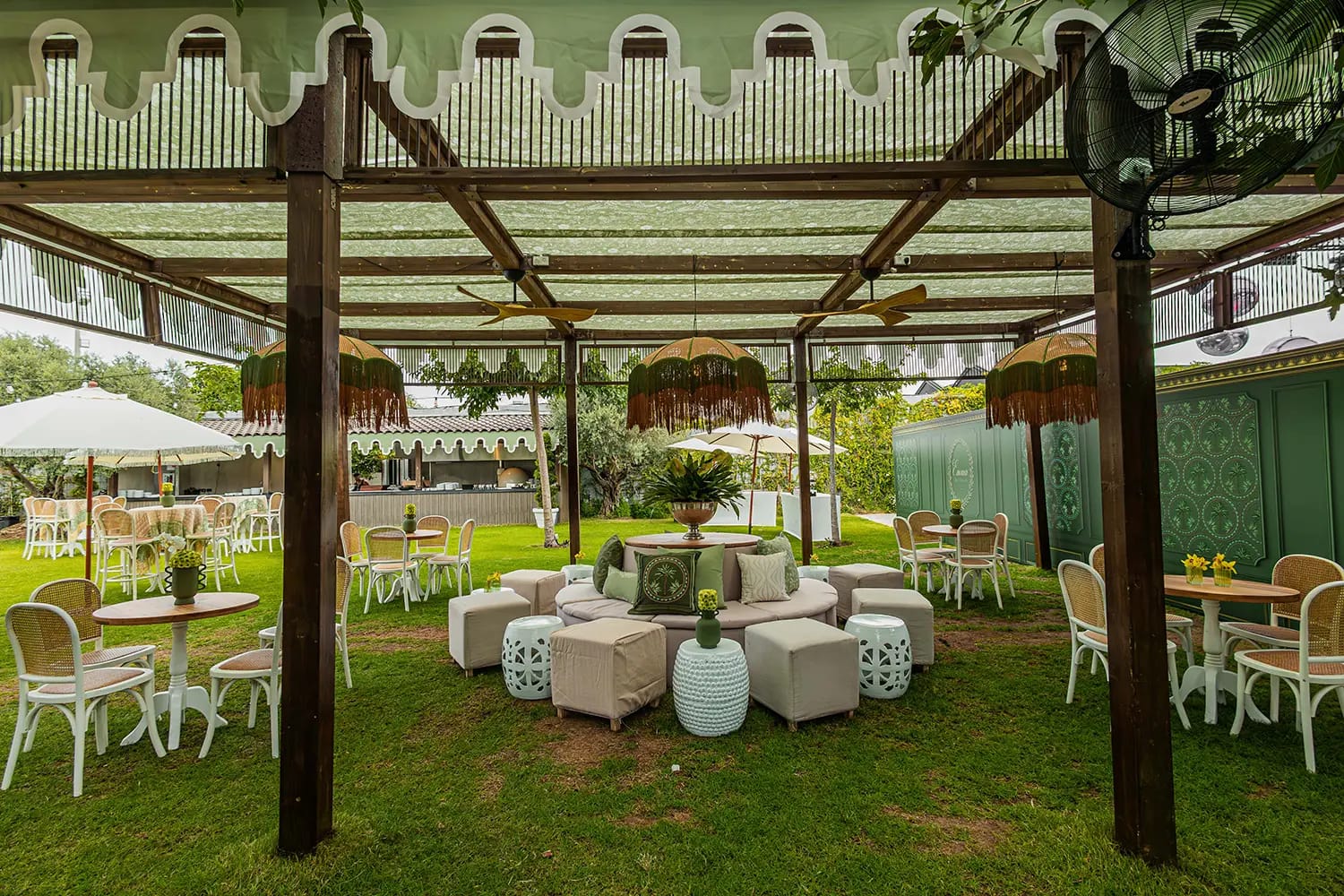 Pergola en bois à auvent festonné et abat-jour à franges au-dessus d'un salon sur pelouse, chaises blanches et poufs