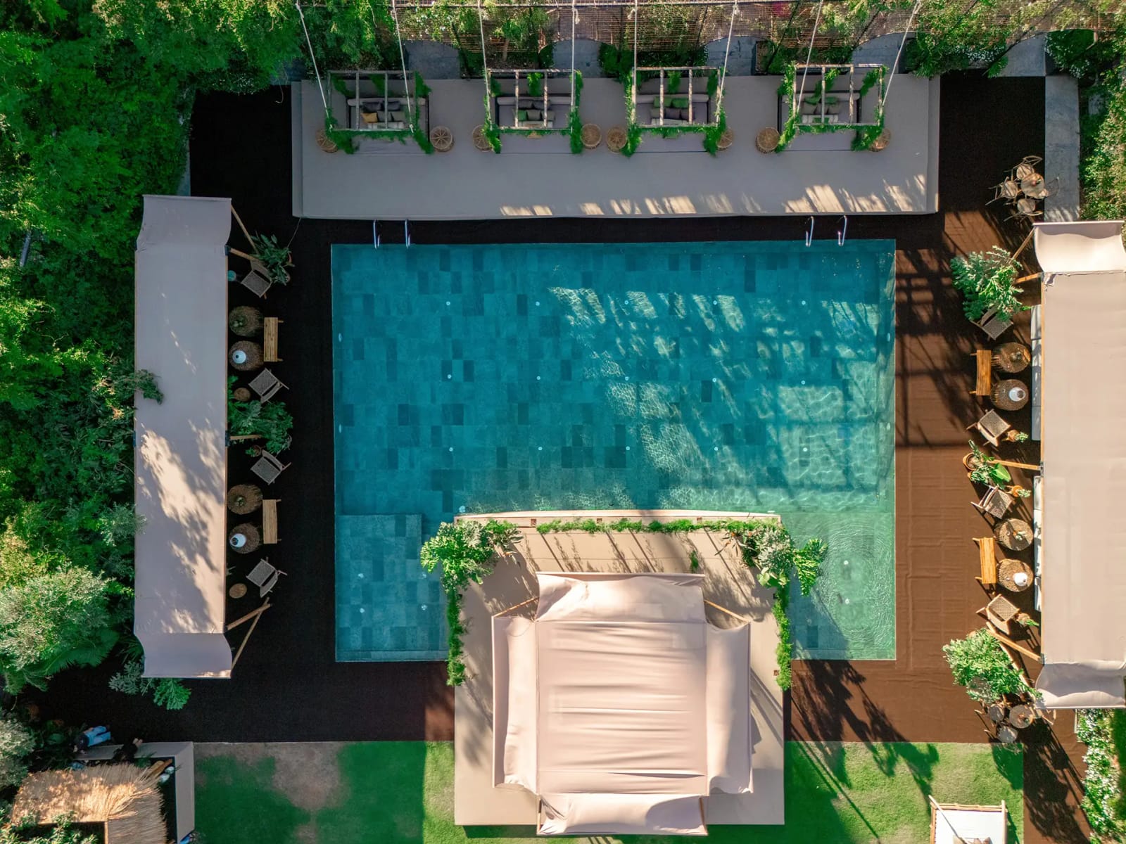 Drone view of a square turquoise pool ringed by a dark deck, lounge seating and a pale canopied daybed