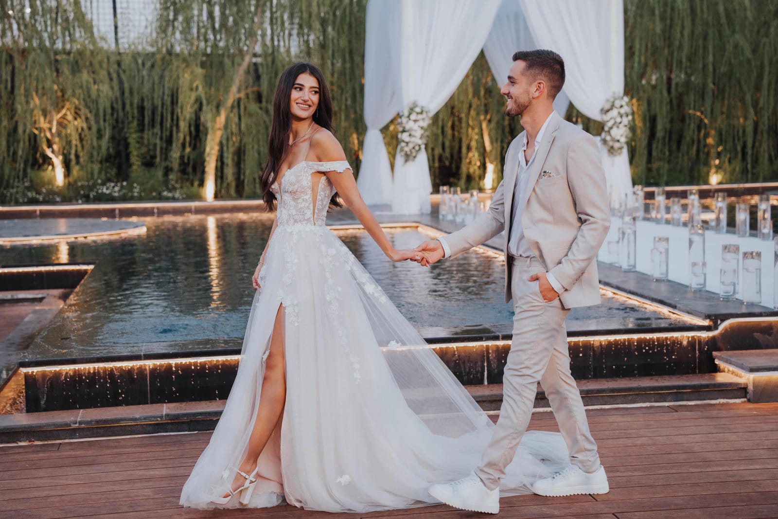A couple walking hand in hand beside a lit pool before white drapes and willow trees, he in a light suit, she in a tulle gown