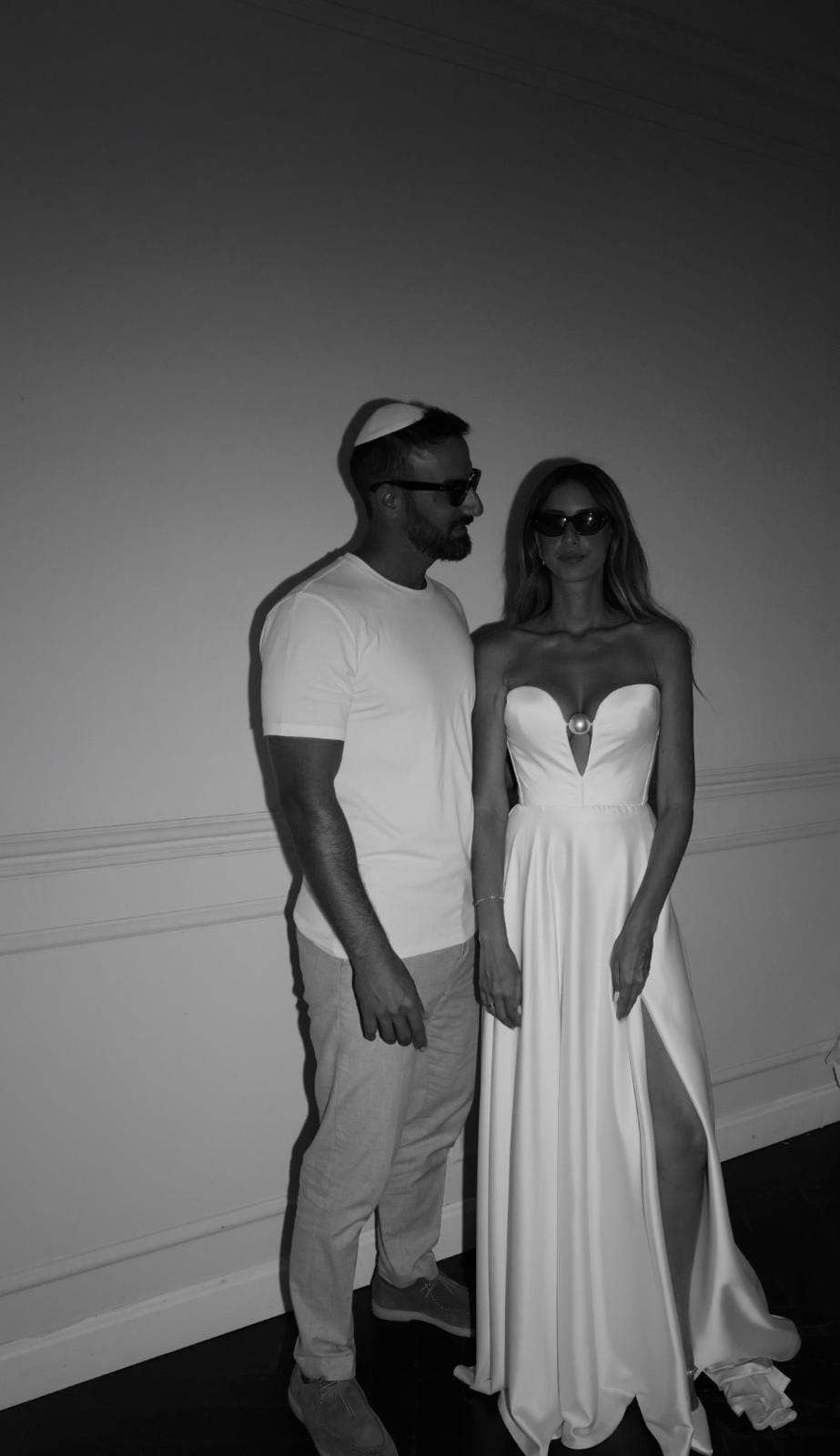 Black-and-white portrait of a couple in a plain room, he in a white T-shirt and kippah, she in a strapless white gown