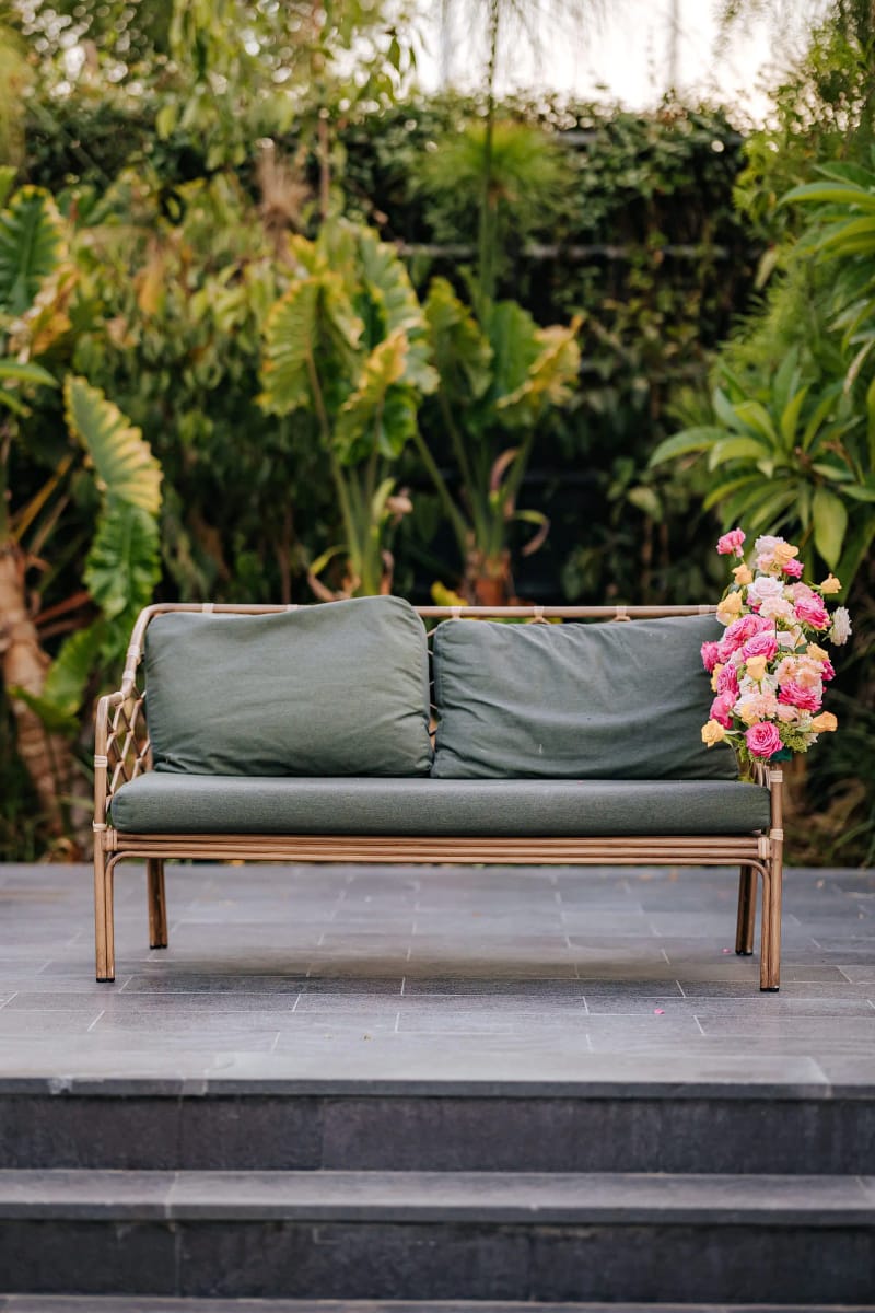 Canapé vert avec une composition de fleurs roses sur le côté, sur une terrasse en pierre devant des plantes tropicales