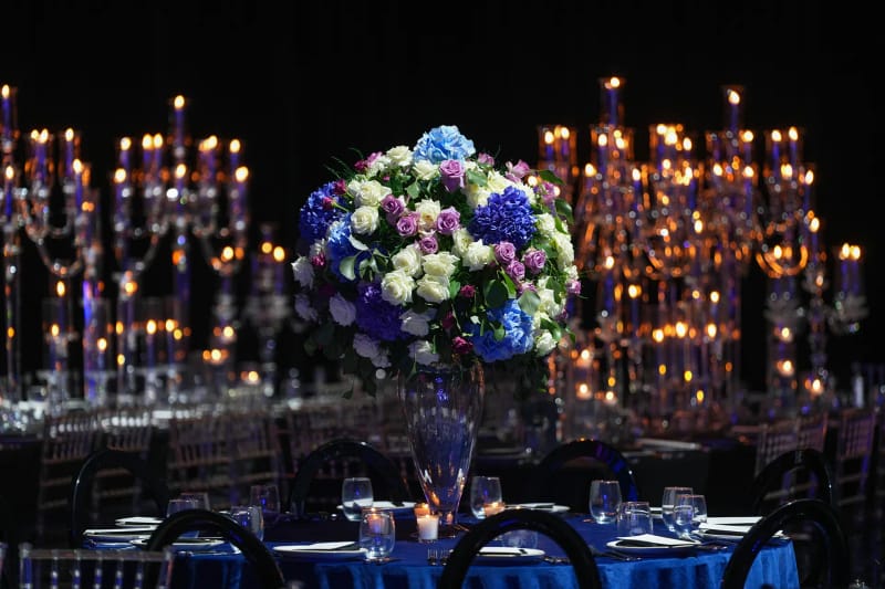 Tall glowing candelabras behind a large bouquet of white, blue and purple flowers on tables with blue cloths