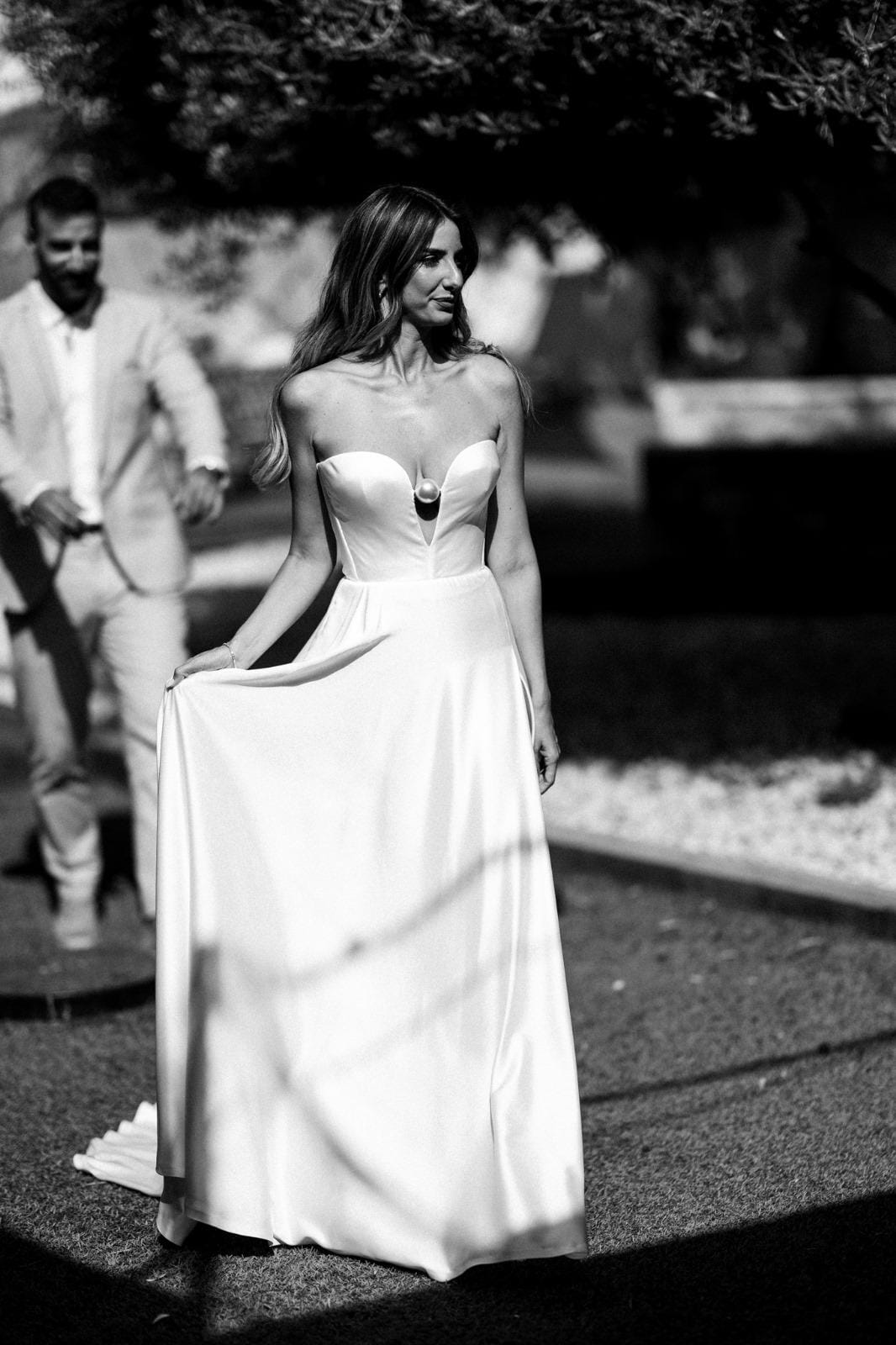 Black-and-white image of a bride lifting her white skirt in a dappled garden with a man standing behind her