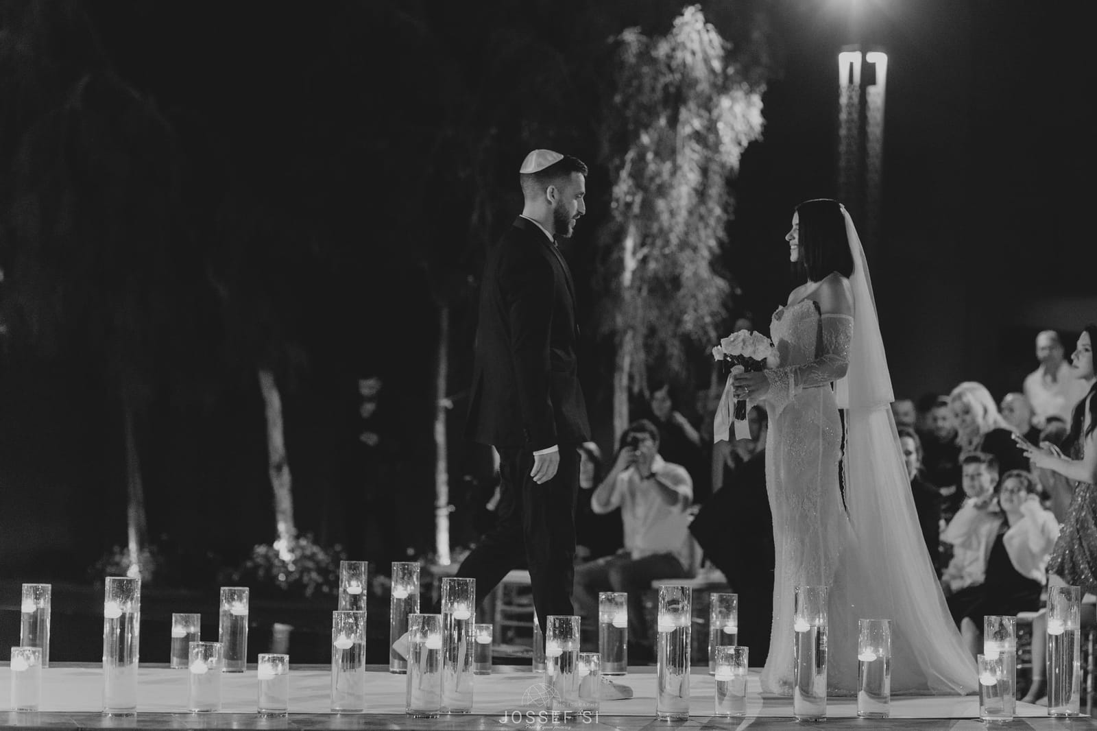 Vue en noir et blanc d'un couple face à face lors d'une cérémonie, invités derrière et photophores en verre devant