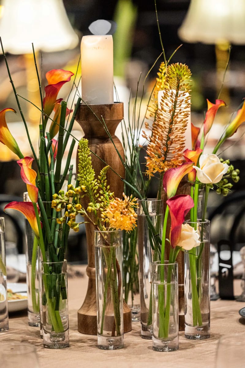 Callas orange et autres hautes fleurs dans des cylindres de verre près d'une bougie sur un bougeoir en bois