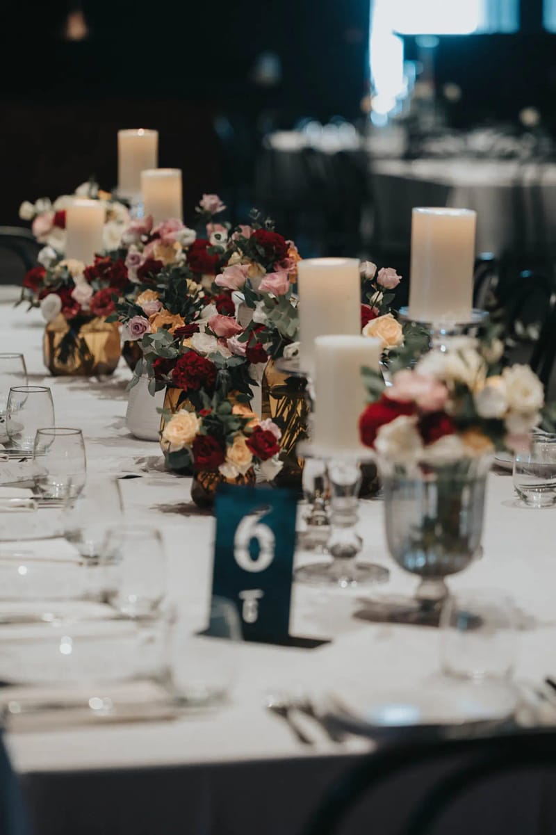 Longue table nappée de blanc avec bougies pilier, fleurs rouges et roses et numéro de table bleu-vert
