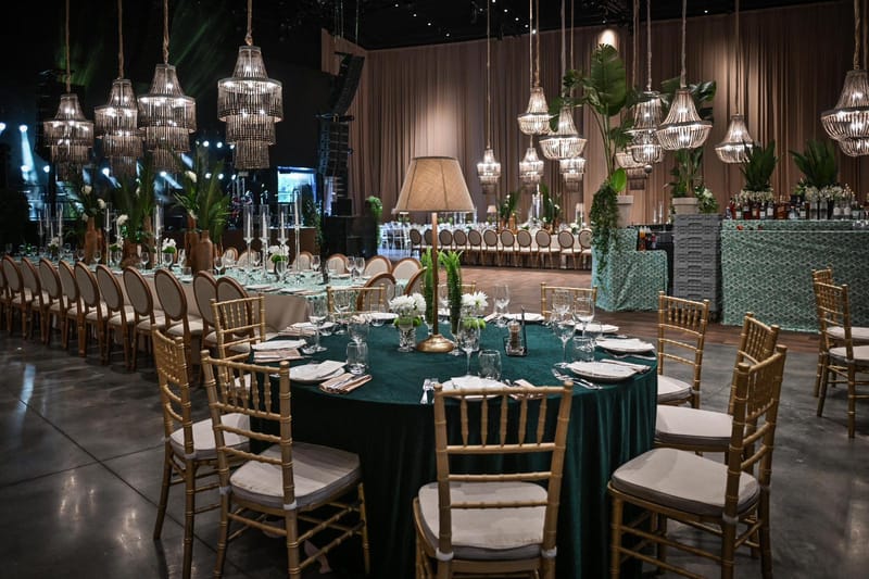Round table with a dark green cloth and gold chairs in a hall with crystal chandeliers, drapes and a green bar behind