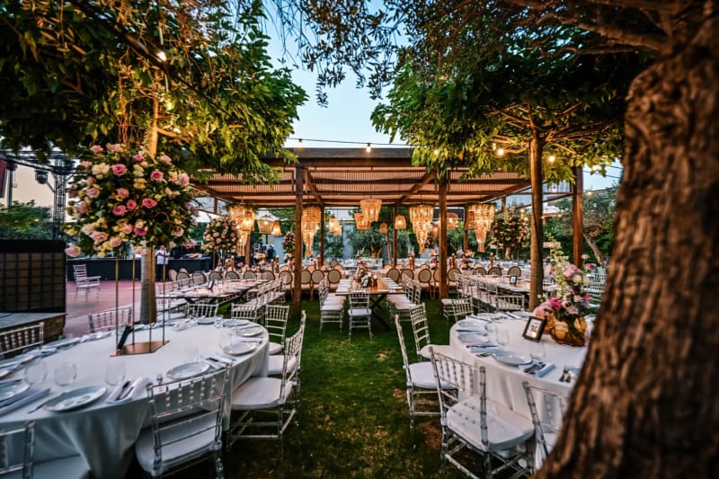 Under a large tree, round tables with a tall pink floral centerpiece and a pergola with hanging chandeliers behind