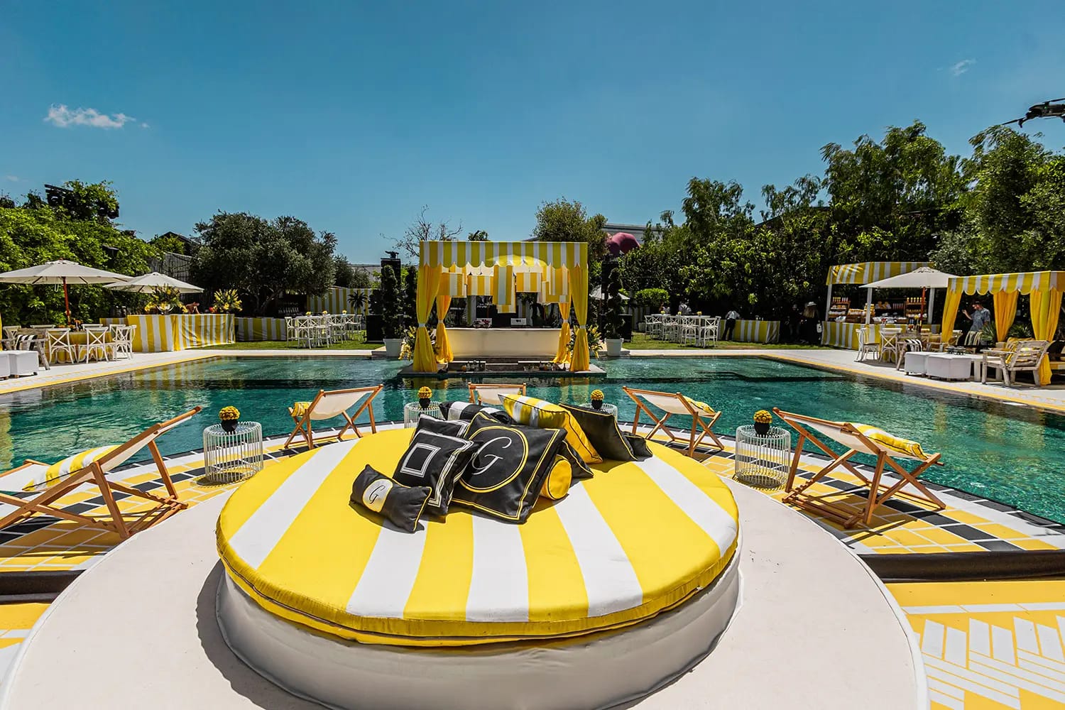 Round striped daybed with black and yellow cushions facing the pool, with deck chairs and a canopy on the far side
