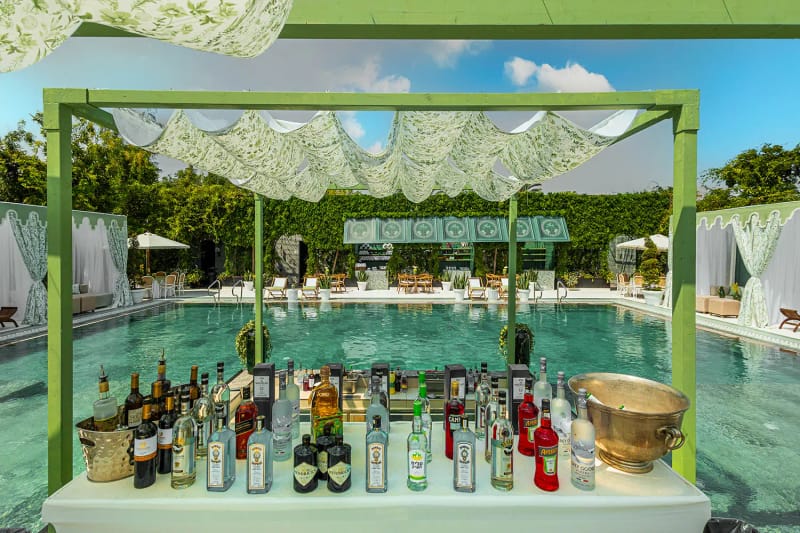 Comptoir de bar avec bouteilles et seau à glace en laiton sous une structure verte drapée de tissu feuillagé, bassin derrière